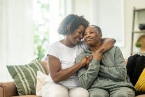 A middle aged woman of African decent wraps her arm around her Mother as she embraces her and they pose for a portrait. They are both dressed comfortably and are laughing as they enjoy their time together.
